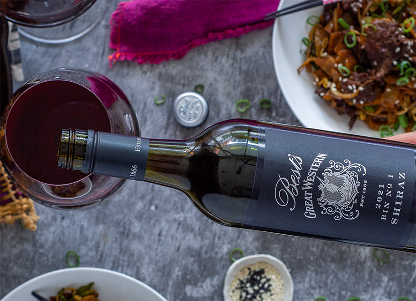 Hand pouring Best's Great Western Bin No. 1 Shiraz into a glass next to a dish of Cantonese beef and noodles.